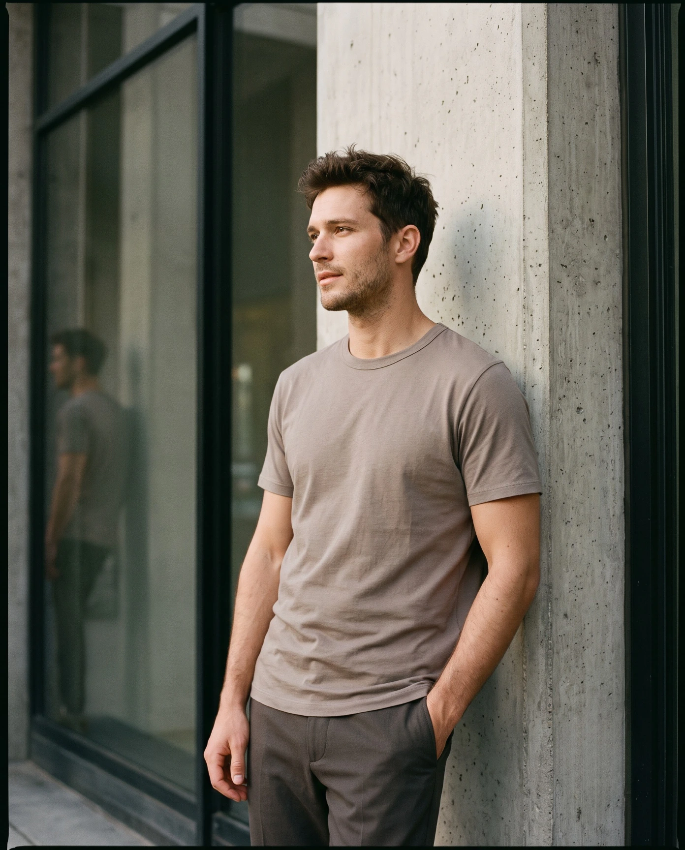 Man wearing a taupe T-shirt and dark relaxed trousers leaning against a concrete building